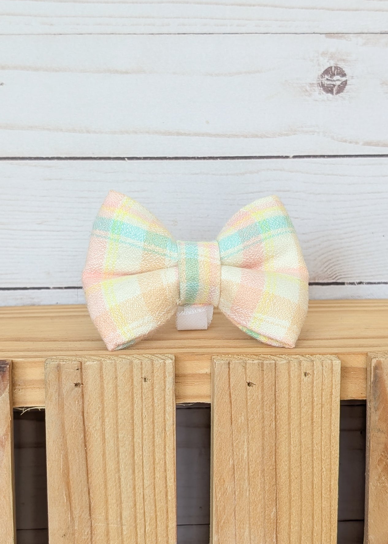 A pastel plaid bow tie with soft yellow, pink, and mint green stripes, resting on a light wooden fence against a white wood plank background.