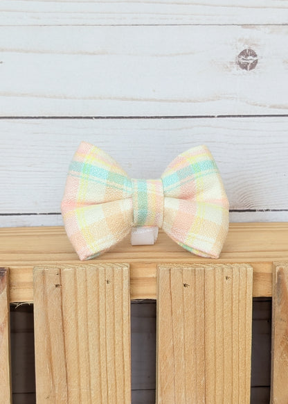 A pastel plaid bow tie with soft yellow, pink, and mint green stripes, resting on a light wooden fence against a white wood plank background.
