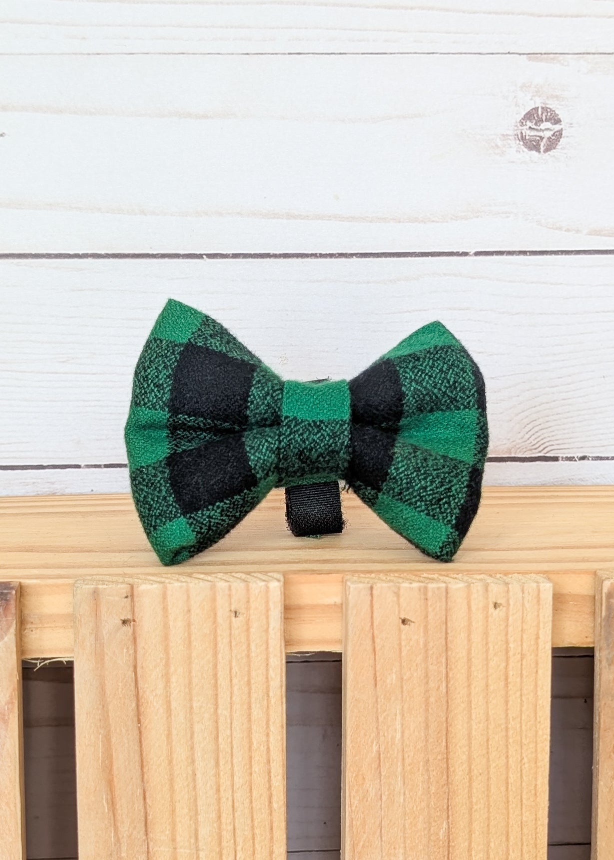 A green and black buffalo plaid bow tie displayed on a light wood slatted surface against a white wood plank background.