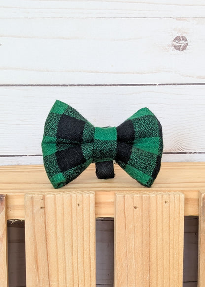 A green and black buffalo plaid bow tie displayed on a light wood slatted surface against a white wood plank background.