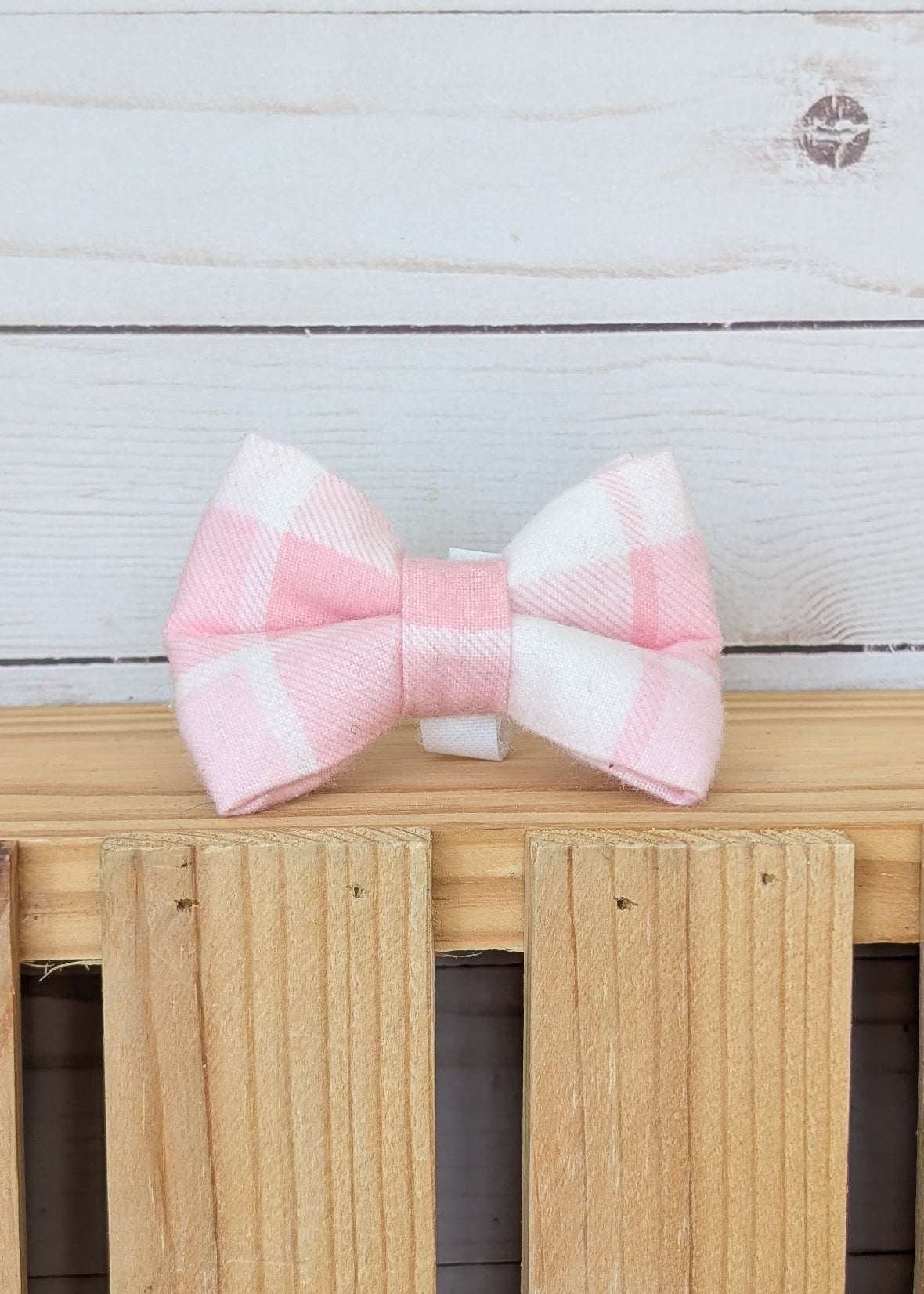 A pink and white gingham bow tie displayed on a light wood slatted surface against a white wood plank background.