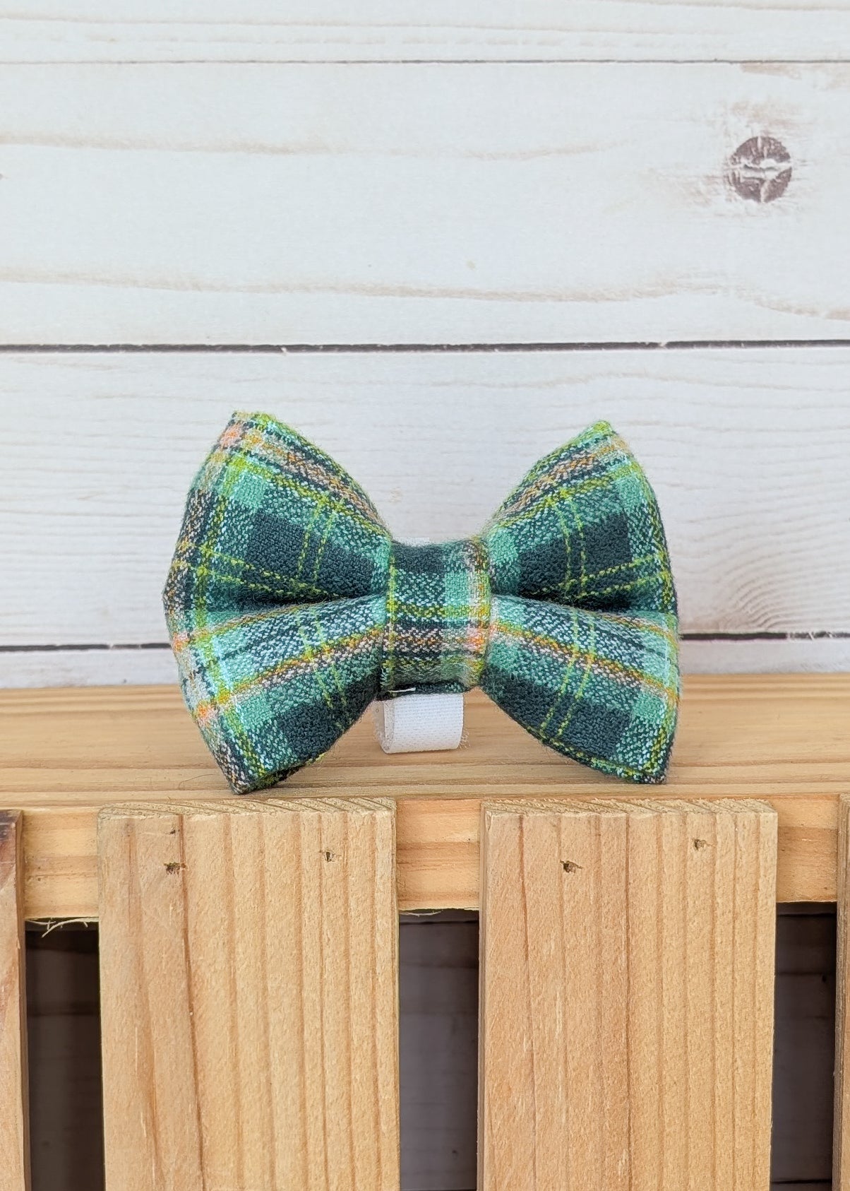 A green and blue tartan plaid bow tie with yellow and white stripes, displayed on a light wood slatted surface against a white wood plank background.