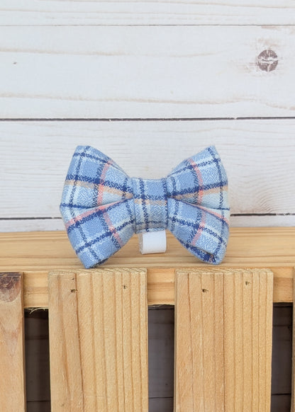 A small blue plaid bow tie with white and orange stripes, displayed on a wooden stand against a white wood plank background.