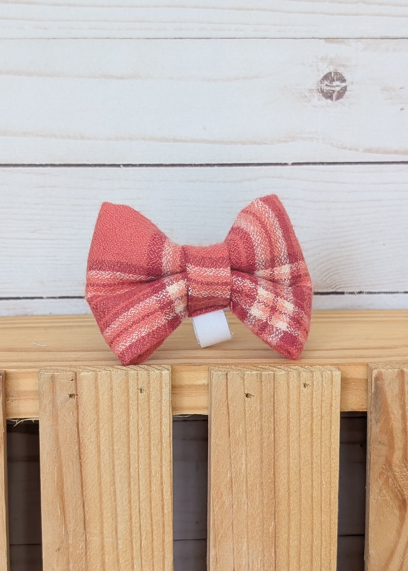 A red and pink plaid bow tie displayed on a light wood slatted surface against a white wood plank background.