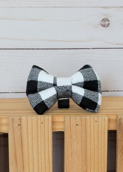 A black and white buffalo check plaid bow tie displayed on a light wood surface against a white wood plank background.