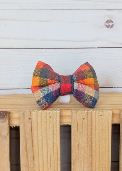 A multicolor plaid bow tie with orange, red, blue, and beige tones, displayed on a light wooden surface against a white wood plank background.
