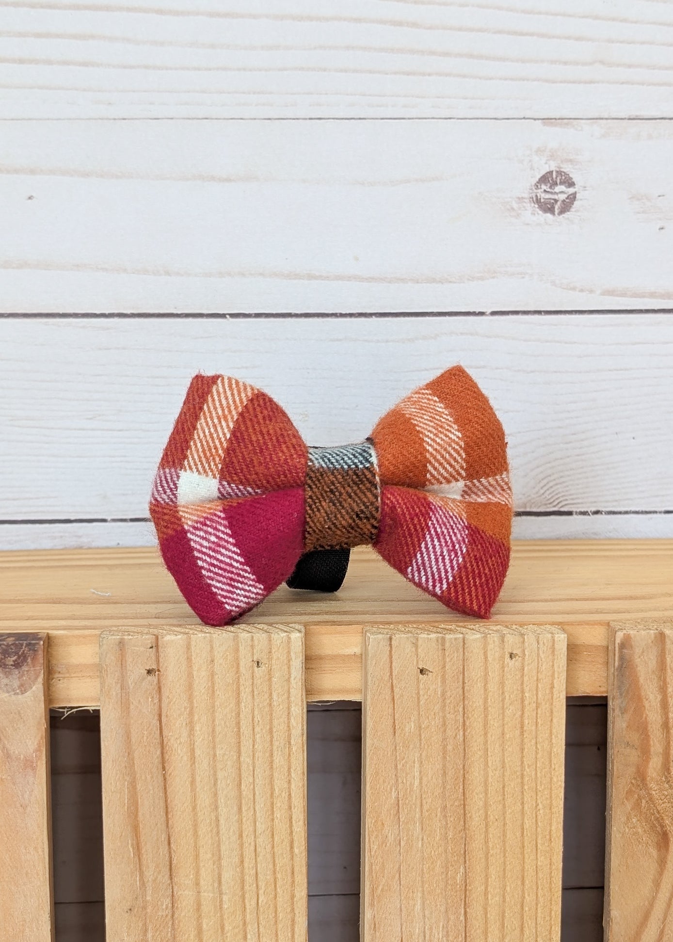A small fabric bow tie with an autumn plaid pattern in orange, red, pink, and white, resting on a light wood slatted surface against a white wood plank background.