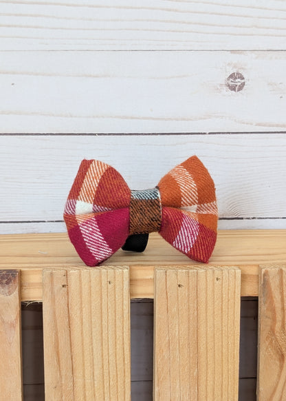 A small fabric bow tie with an autumn plaid pattern in orange, red, pink, and white, resting on a light wood slatted surface against a white wood plank background.