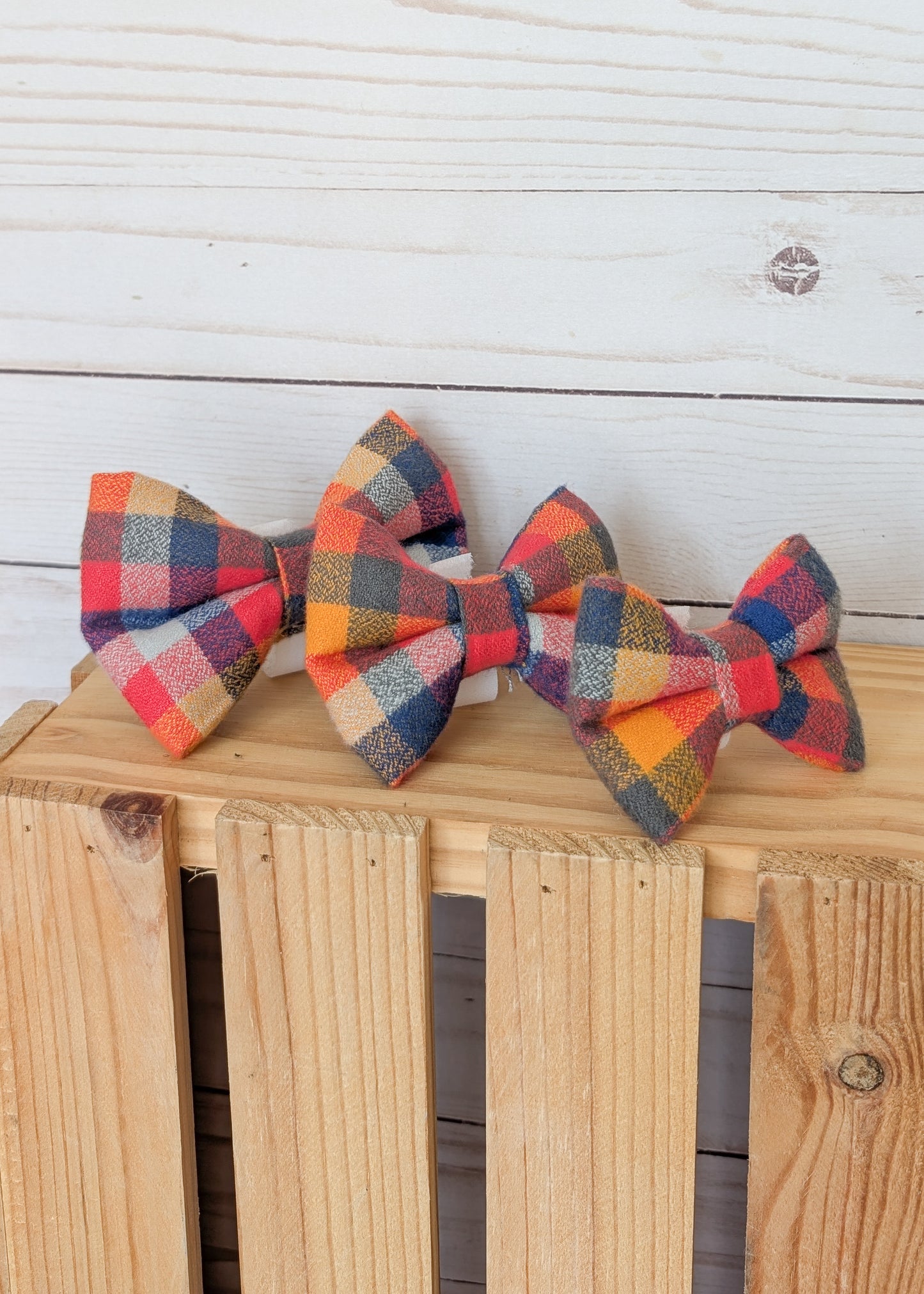 Three fabric bow ties with a multicolor plaid pattern in red, orange, yellow, blue, and beige, displayed on a wooden crate against a white wood plank background.