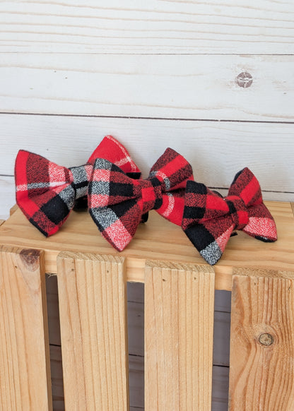 Three red, black, and white plaid bow ties arranged on a wooden crate against a white wood plank background.