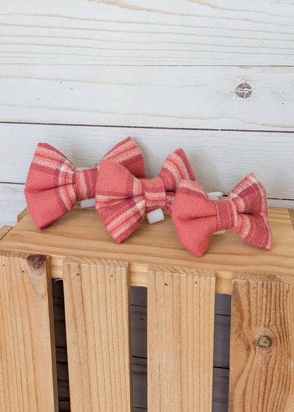 Three small flannel bow ties in a pink and red  plaid pattern, displayed on a wooden crate against a white wood plank background.
