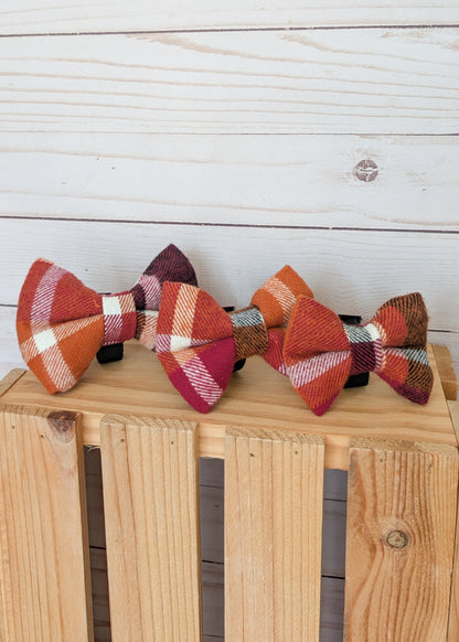 Three small fabric bow ties with autumn plaid patterns in orange, rust, burgundy, cream, and brown, displayed on a wooden crate against a white wood plank background.