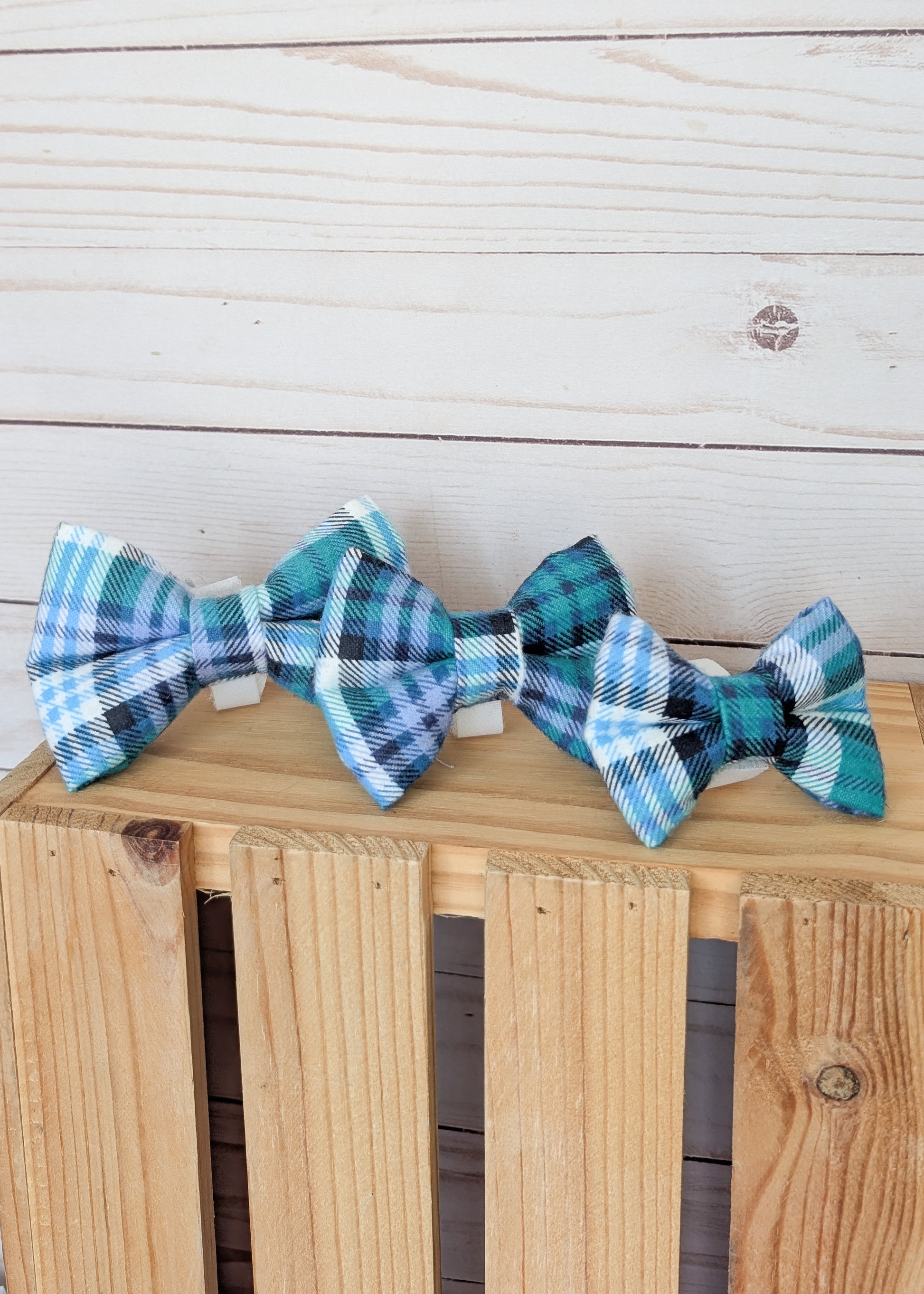 Three small plaid bow ties in shades of teal, blue, white, and black, displayed on a wooden crate against a light wood plank background.