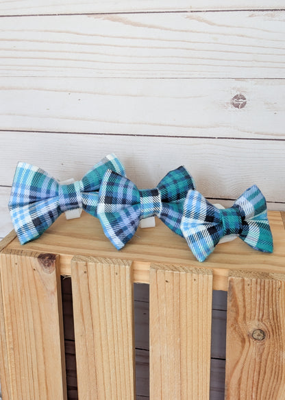 Three small plaid bow ties in shades of teal, blue, white, and black, displayed on a wooden crate against a light wood plank background.