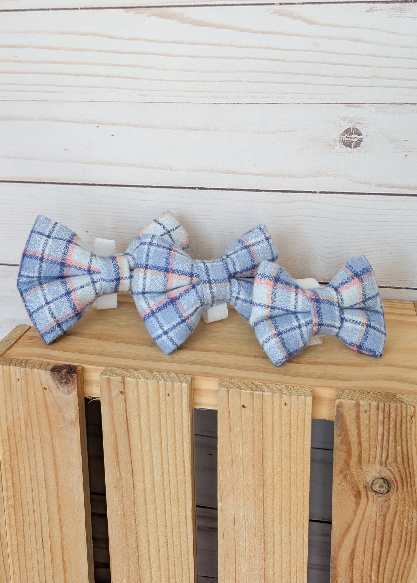 Three small fabric bow ties with a light blue, white, and pink plaid pattern arranged on a wooden crate against a white wood plank background.