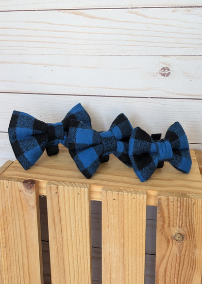 Three matching blue and black buffalo plaid bow ties displayed on a light wood crate against a white wood plank background.