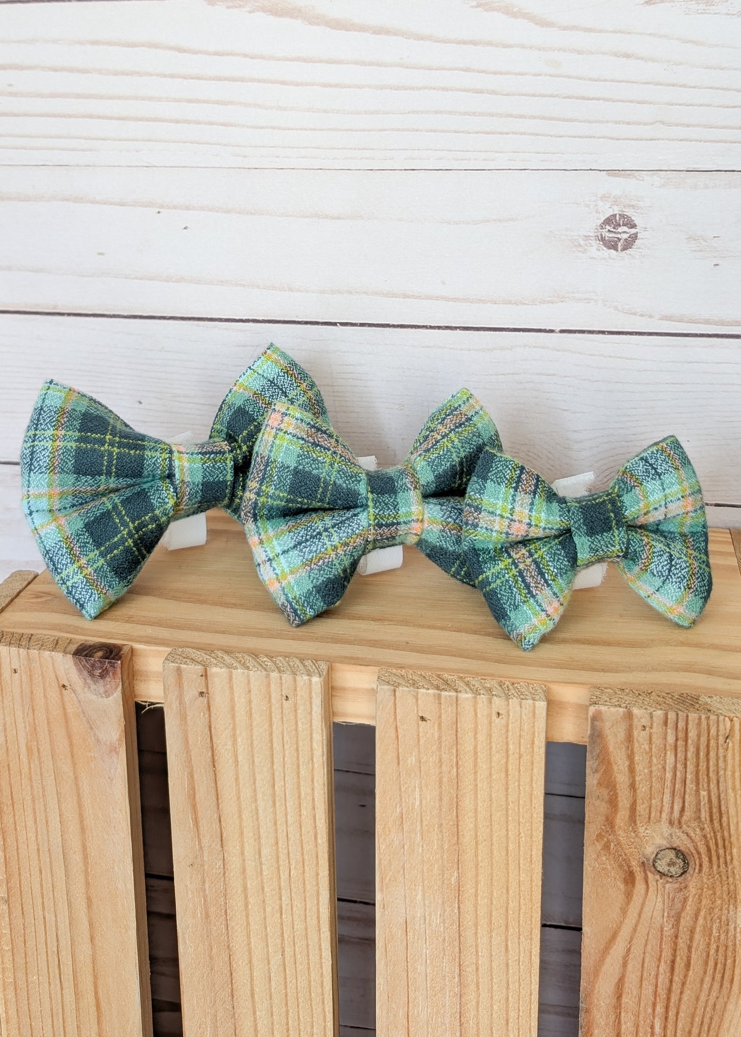 Three small plaid bow ties in green, blue, yellow, and white tartan patterns, displayed on a light wood crate against a white wood plank background.