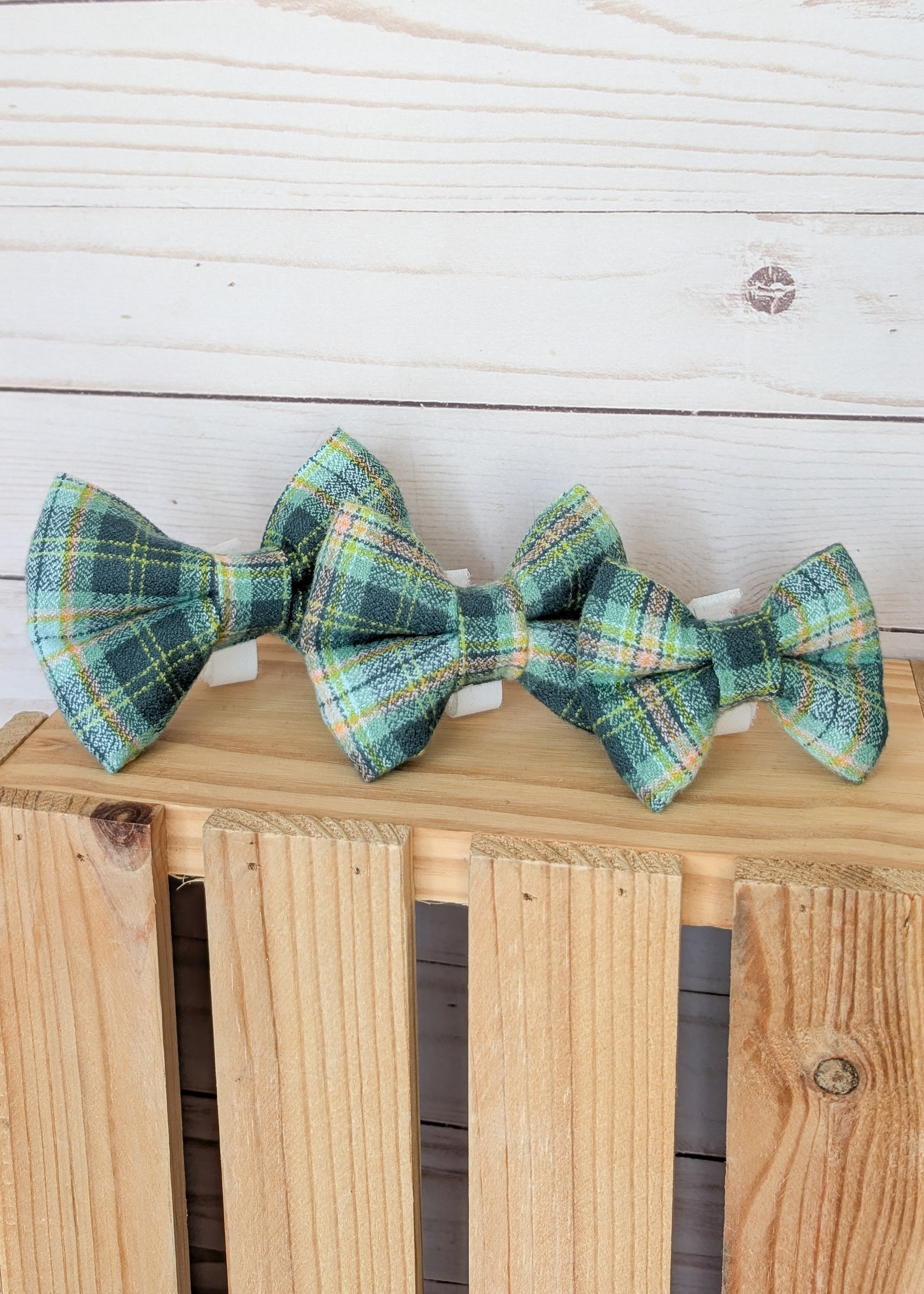 Three small plaid bow ties in green, blue, yellow, and white tartan patterns, displayed on a light wood crate against a white wood plank background.