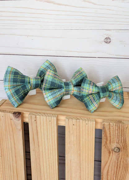 Three small plaid bow ties in green, blue, yellow, and white tartan patterns, displayed on a light wood crate against a white wood plank background.