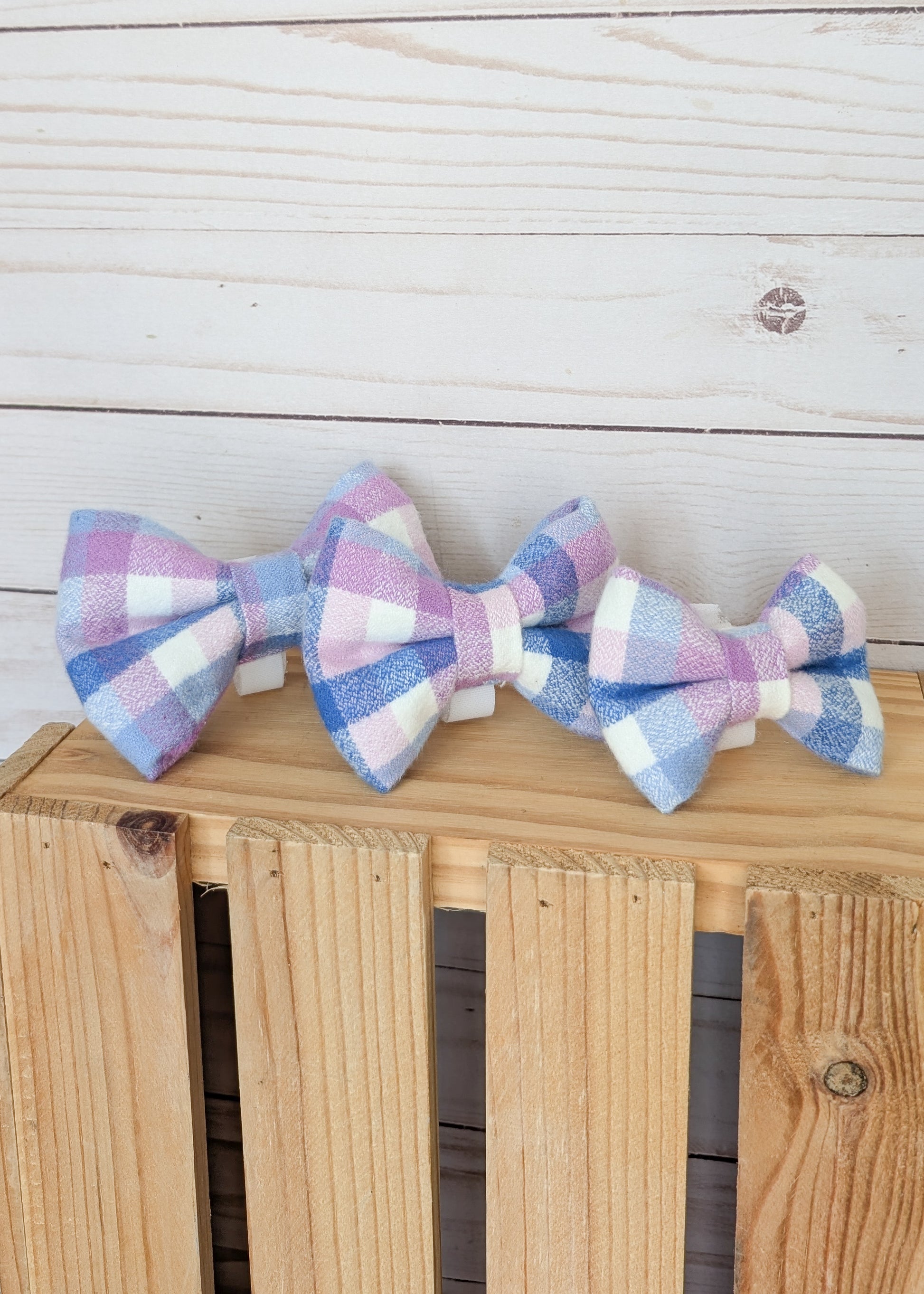 Three fabric bow ties with a pastel gingham plaid pattern in pink, purple, blue, and white, displayed on a wooden crate against a white wood plank background.
