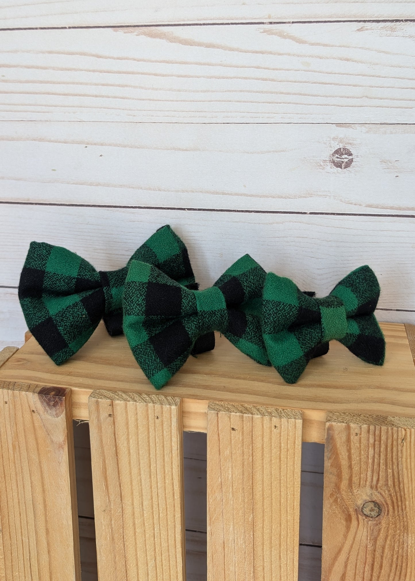 Two green and black buffalo plaid bow ties displayed on a wooden crate against a white wood plank background.