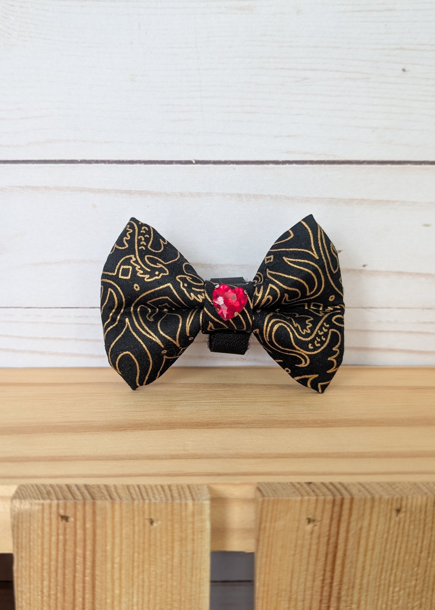 A black and gold patterned bow tie with a small red heart shaped crystal accent in the center, displayed on a light wooden surface against a white wood plank background.