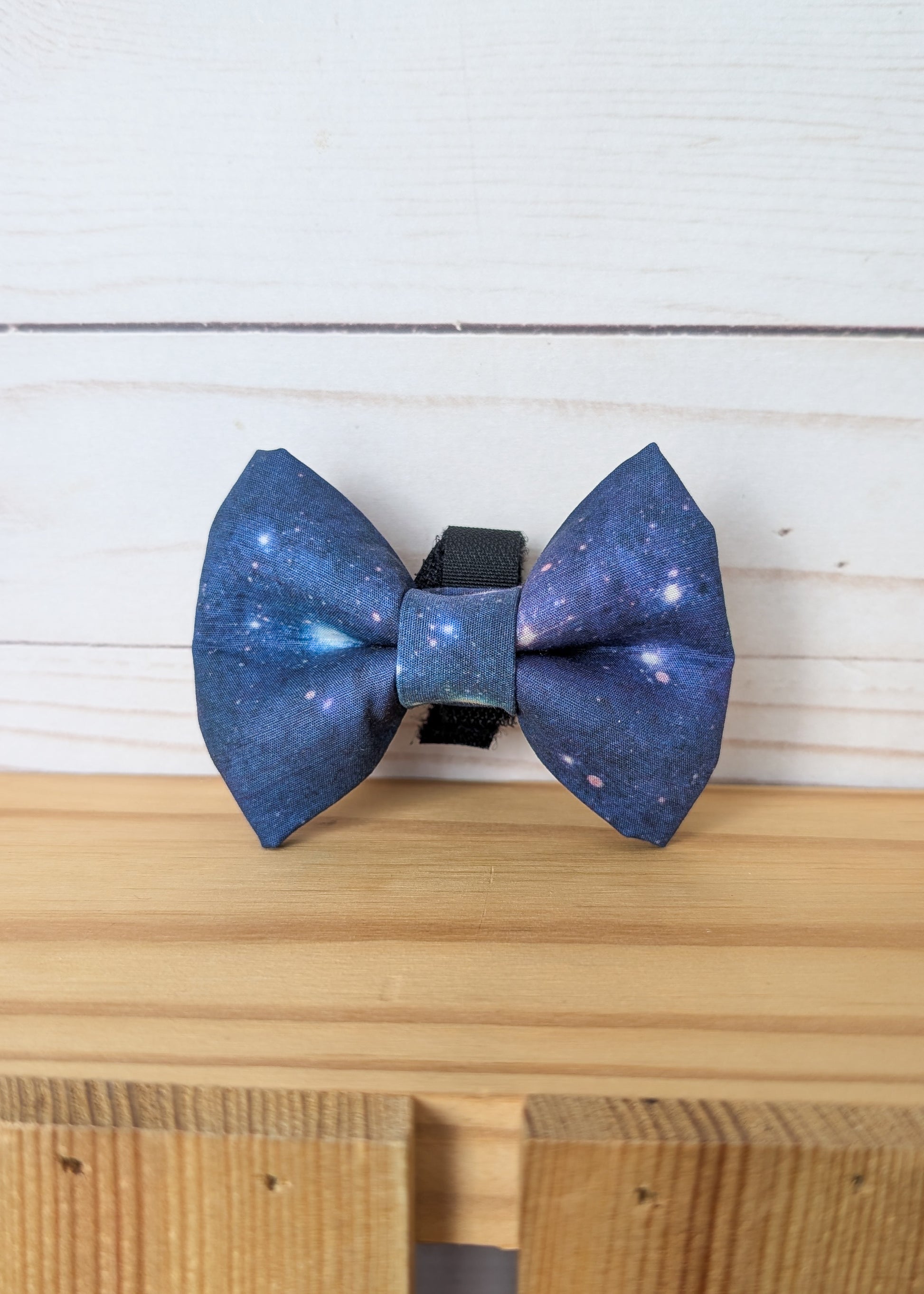 A galaxy-printed bow tie with a blue and purple cosmic pattern, resting on a light wooden surface against a white wood plank background.