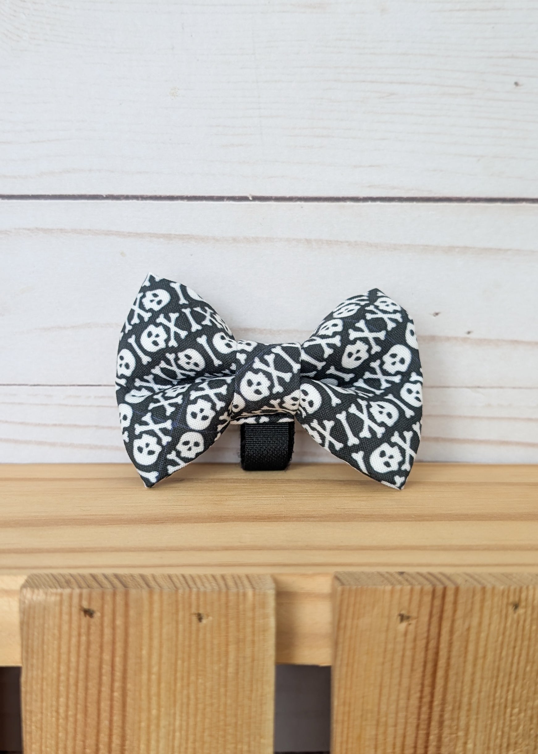 A black and white skull and crossbones patterned bow tie displayed on a light wooden surface against a white wood plank background.