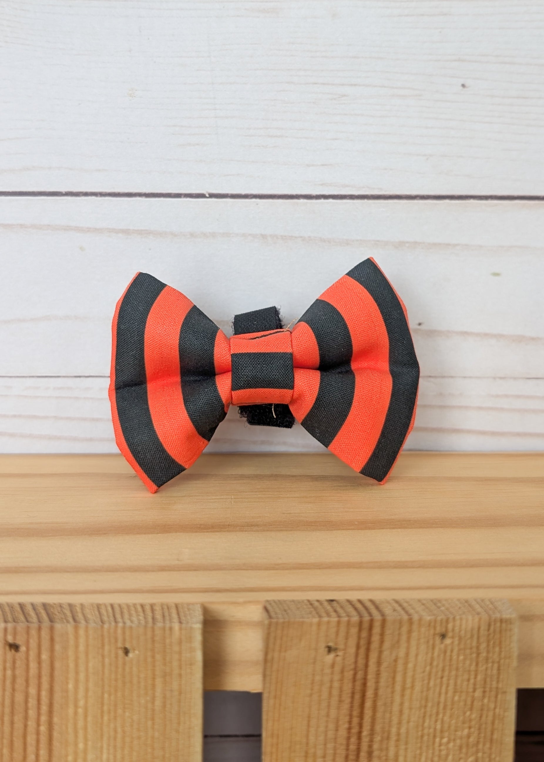 A small striped bow tie in bright orange and black, displayed on a light wood surface against a white wood plank background.