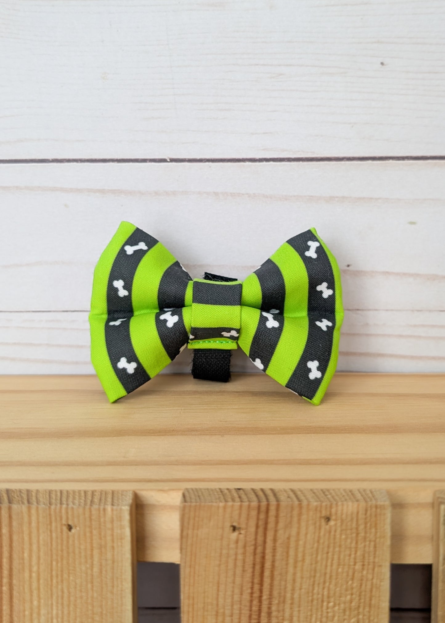 A pet bow tie with lime green and black stripes and small white bone patterns, displayed on a wooden surface against a white wood plank background.