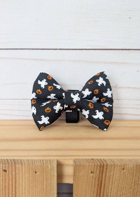 A black bow tie with a white ghost and orange pumpkin pattern, displayed on a wooden surface against a white wood plank background.