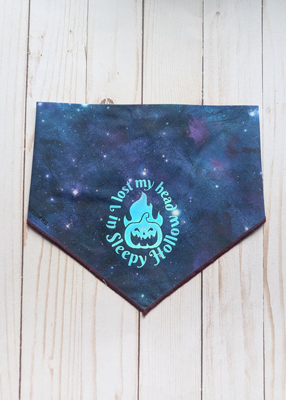 A dog bandana with a dark blue and purple galaxy print, featuring a light blue graphic of a jack-o'-lantern with a flame and the text 'I lost my head in Sleepy Hollow'. The bandana is laid flat on a light-colored wooden plank background.