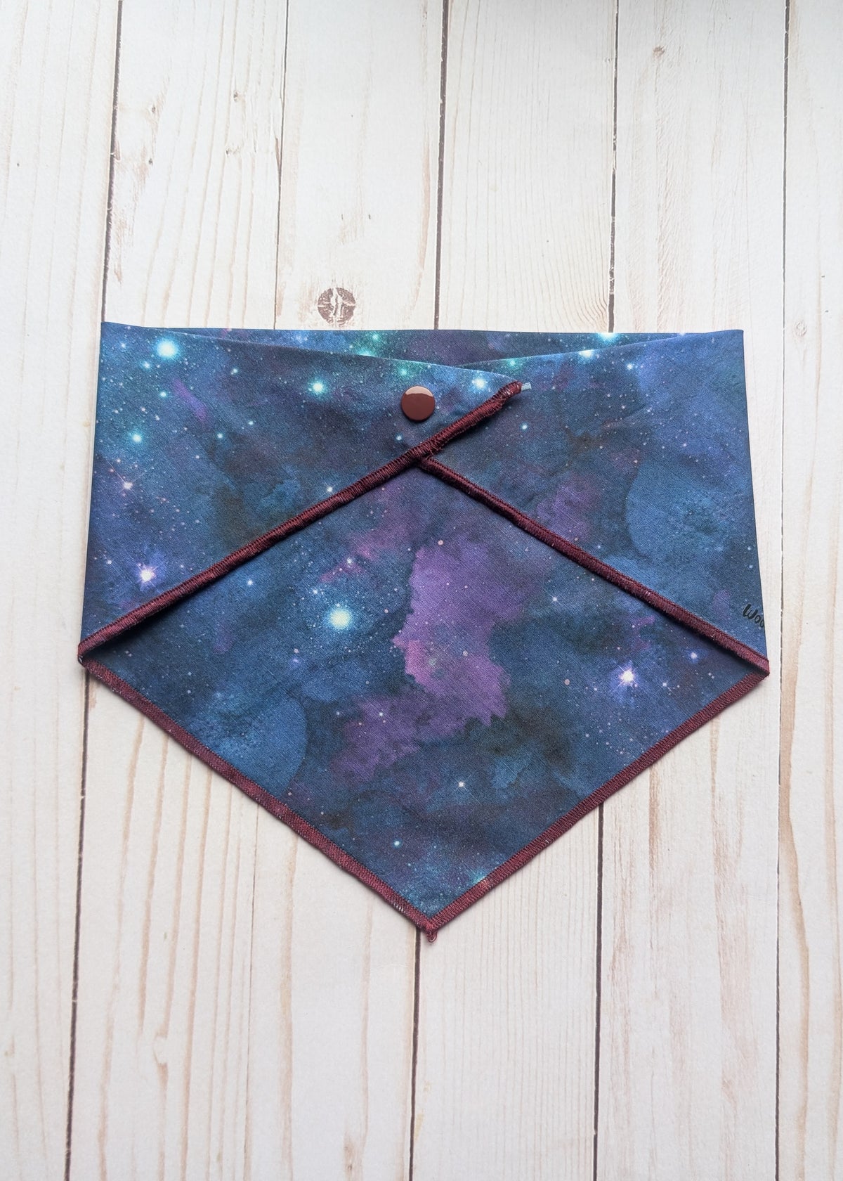 A diamond-shaped bandana with a galaxy print in shades of blue, purple, and black, featuring a dark red stitched border and a snap-button closure, laid flat on a light wood plank background.