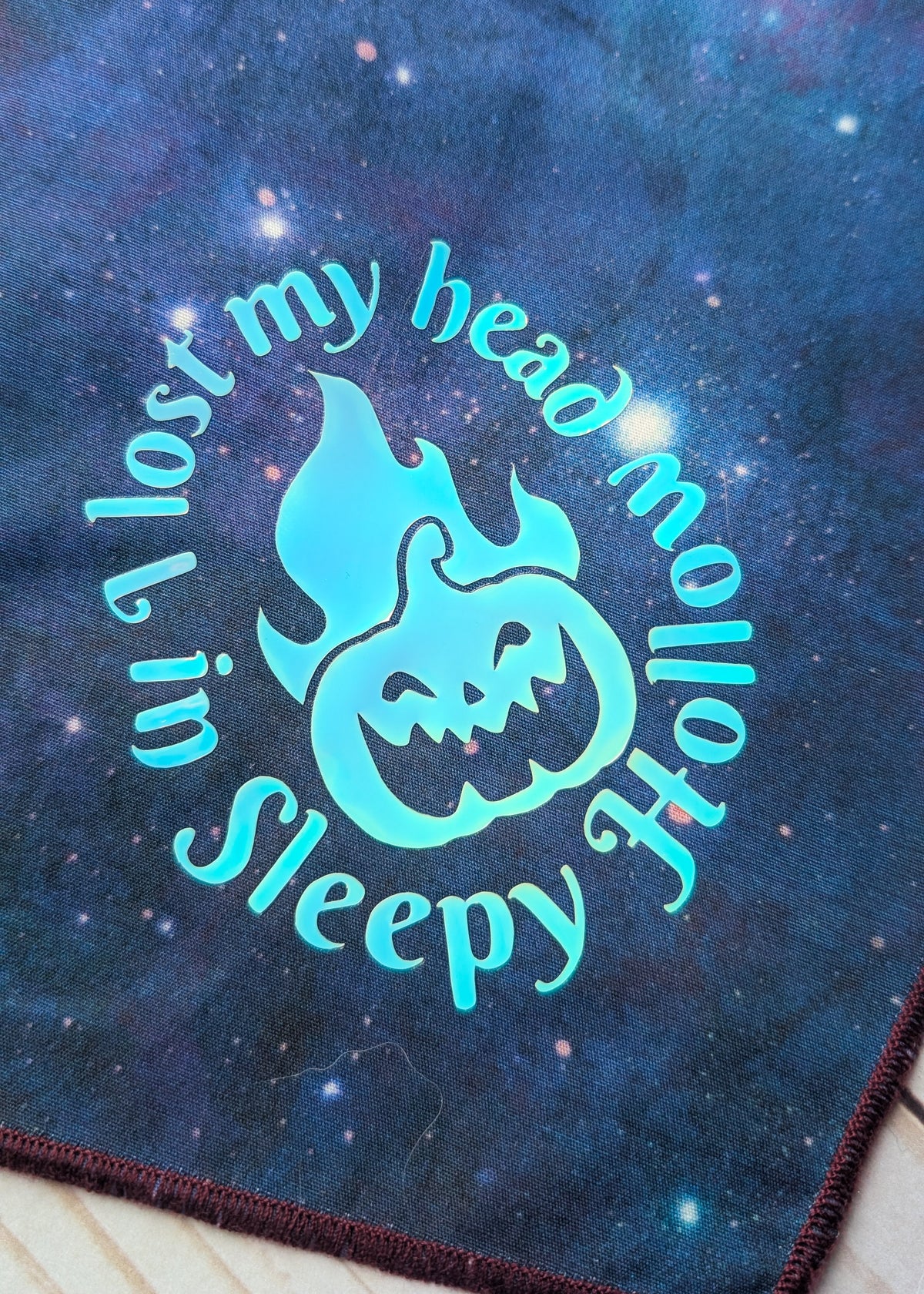 A close-up of a dark blue galaxy-patterned fabric featuring a glowing turquoise graphic of a jack-o'-lantern with flames and the text 'I lost my head in Sleepy Hollow'