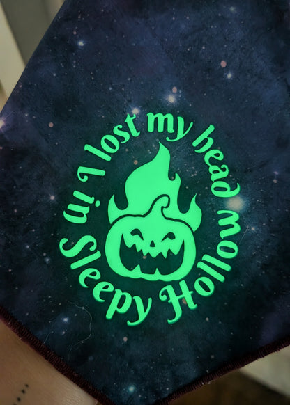A close-up of a dark galaxy-print bandana or scarf featuring a glowing neon green graphic of a flaming jack-o'-lantern with the text 'I lost my head in Sleepy Hollow' curved around it.