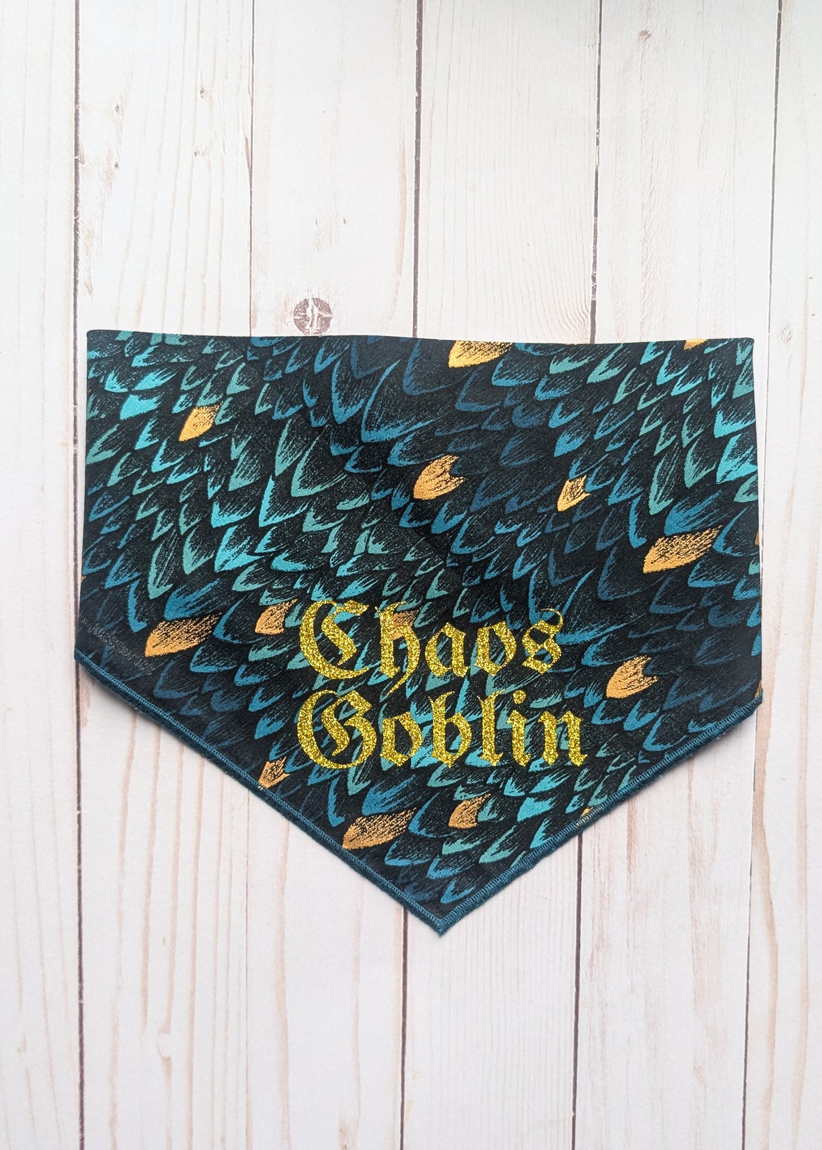 A triangular pet bandana featuring a dark teal and black scale pattern with gold glitter accents and the words 'Chaos Goblin' in gold gothic lettering, laid flat on a white wood plank background.