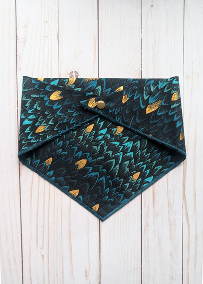 A triangular bandana, teal and gold feather pattern, and a snap button closure, laid flat on a white wood plank surface.