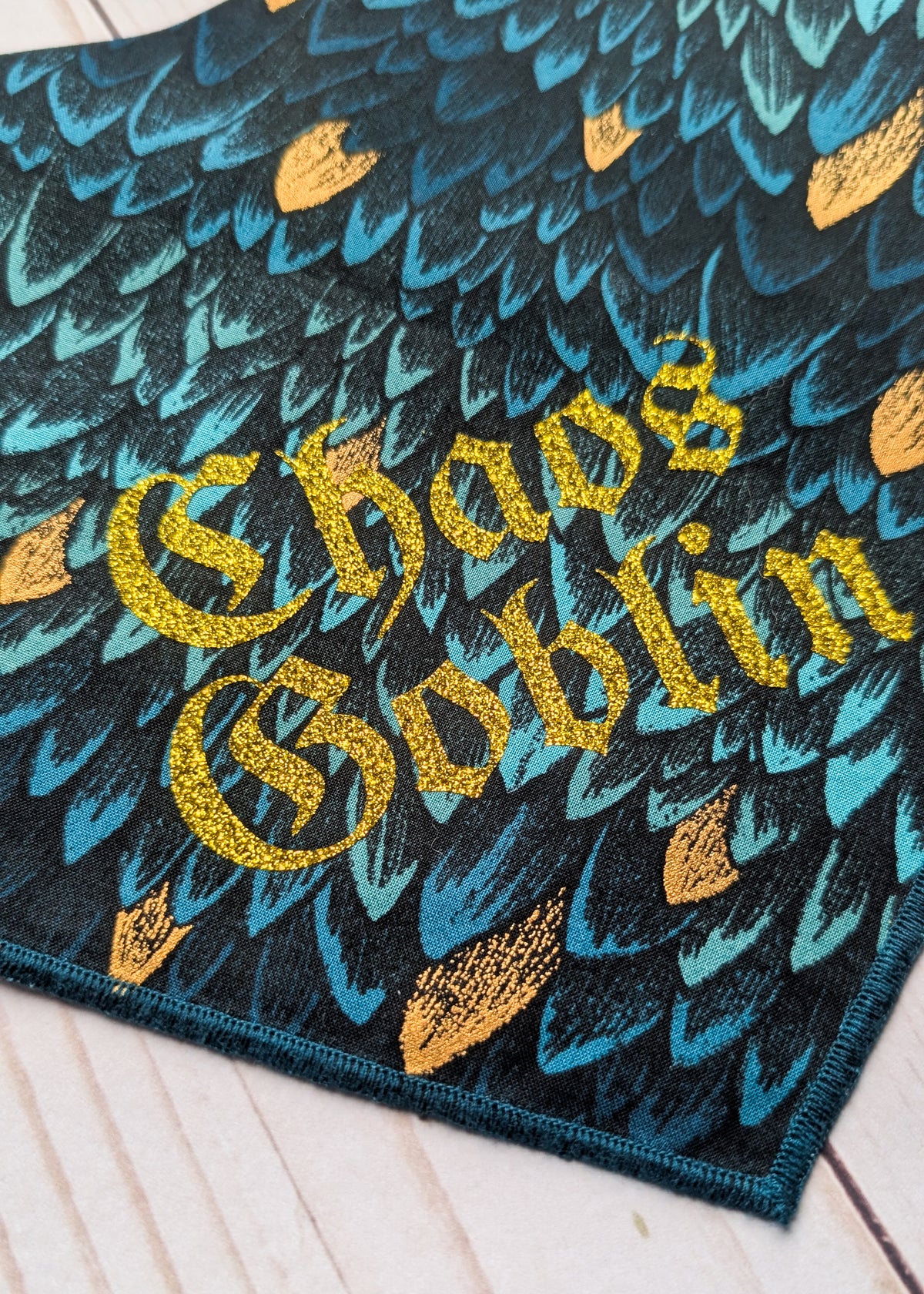 A close-up of a bandana, featuring a black background with a teal and blue feather-like pattern and gold glittery text reading 'Chaos Goblin'.