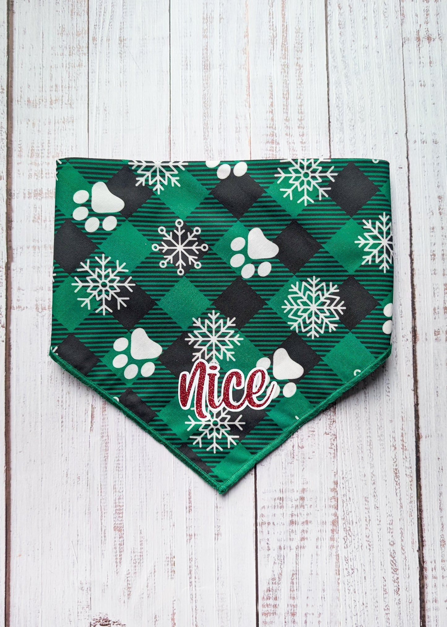 Green and black buffalo plaid bandana with green hem edge, white snowflake and paw print pattern and "nice" as a central text in red shimmer with white outline
