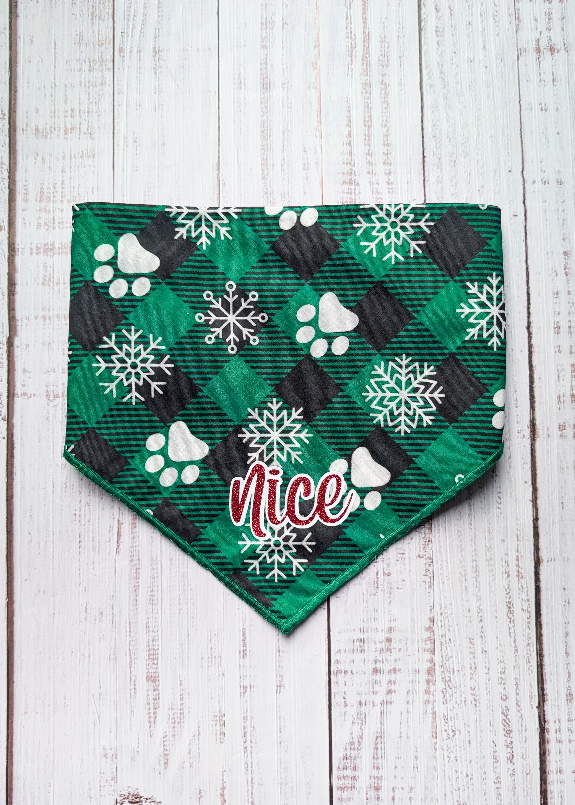 Green and black buffalo plaid bandana with green hem edge, white snowflake and paw print pattern and "nice" as a central text in red shimmer with white outline
